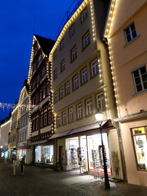 Eine Kopfsteinpflasterstraße in Rothenburg ob der Tauber, Deutschland, geschmückt mit festlichen Weihnachtslichtern, mit Menschen, die spazieren gehen und einem Fahrrad in der Nähe, flankiert von Gebäuden mit Fenstern unter einem sichtbaren Himmel.