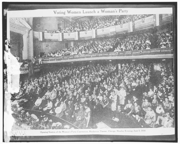 Schwarzes und weißes Foto einer großen Menschenmenge in einem Gebäude mit Schildern, einige sitzen und einige stehen, und Text unten lesend 'Wahlberechtigte Frauen gründen eine Frauenpartei.'