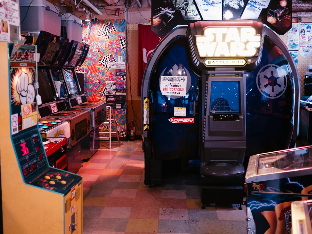 Ein hell erleuchteter Arcade-Raum mit mehreren Maschinen, darunter ein Star Wars Battle Pod, farbige Poster an den Wänden und verstreute Gegenstände, die eine lebendige Atmosphäre schaffen.