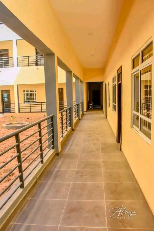 Ein mehrstöckiges Gebäude mit Fenstern, Türen, Säulen und Geländern, das einen langen gefliesten Flur und einen Balkon zeigt, vor anderen Gebäuden unter einem klaren blauen Himmel, mit Miettext unten.