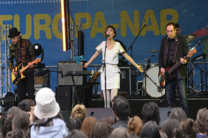Eine Frau in einem weißen Kleid singt auf der Bühne bei einem Konzert, flankiert von zwei Gitarristen, mit einer Menge davor und Lautsprechern im Hintergrund.