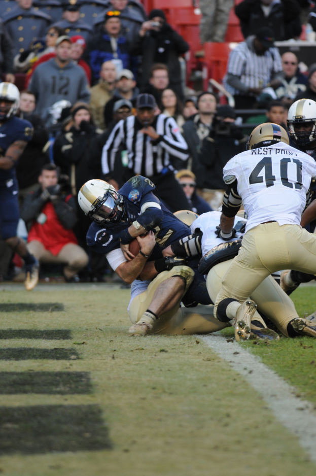Ein Fußballer wird von einer Gruppe von Verteidigern auf einem Rasenfeld getackelt, mit Zuschauern und Kameramännern im Hintergrund, alle Spieler tragen Helme.