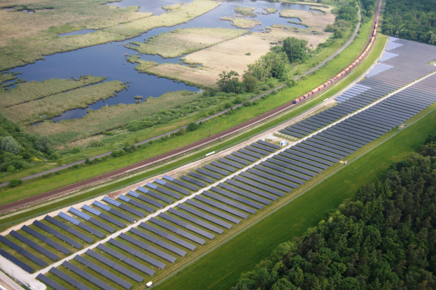 Luftaufnahme einer Solar-Farm mit Panelen in einem Feld, umgeben von Bäumen, Gras, Pflanzen und Wasser, mit einem Zug auf einer nahen Bahnstrecke.
