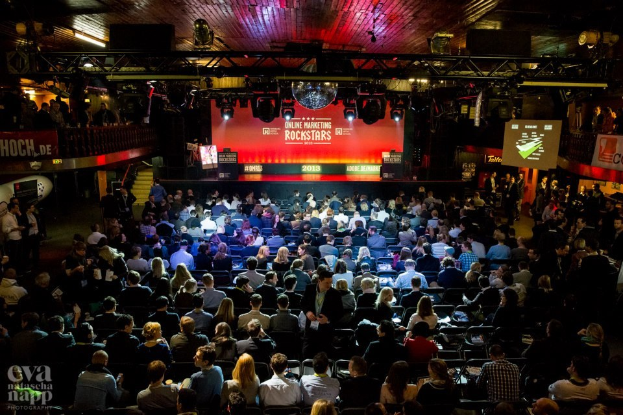 Eine große Zuschauermenge sitzt in Reihen vor einer hell erleuchteten Bühne mit Bildschirmen und Bannern und wartet auf ein Ereignis.