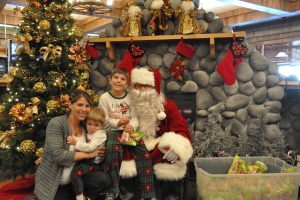 Eine Frau, Kinder und ein Mann in Santa-Klaus-Kostüm mit Brille und Handschuhen stehen neben einem geschmückten Weihnachtsbaum, einer Wand mit Felsen, Spielzeugen, Dekorationen und einem Korb mit Gegenständen, mit Lichtern im Hintergrund.