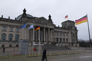 Eine Person geht vor dem Reichstagsgebäude in Berlin, Deutschland, vorbei, das Fenster, Säulen, Bögen, Statuen und umgebende Flaggen mit Stangen, Treppen, Geländern, Bäumen und einem klaren blauen Himmel im Hintergrund zeigt.