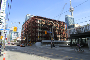 Eine belebte Stadtstraße mit einem großen Gebäude im Hintergrund, Fahrzeugen, einem Radfahrer, Passanten, Verkehrsampeln, Pfählen mit Schildern, einem Kran und dem Himmel.