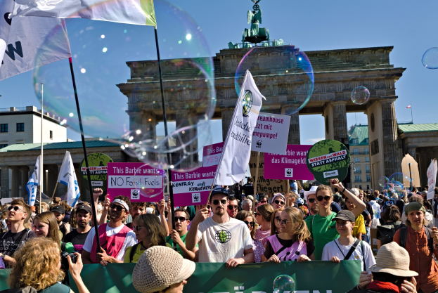 Eine Menschenmenge vor dem Brandenburger Tor in Berlin, viele mit Mützen und Schutzbrillen, einige mit Fahnen und Transparenten mit Text, im Hintergrund Gebäude, ein von einer Statue gekrönter Bogen und ein mit Seifenblasen gefüllter Himmel.