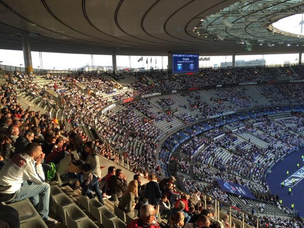 Großes Publikum in einem Stadion bei einem Fußballspiel mit einer Bühne, Fahnen und einem Bildschirm im Hintergrund, identifiziert als Allianz Arena in München, Deutschland.