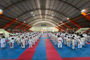 Eine große Gruppe von Menschen auf einer blauen und roten Matte in einer Turnhalle, mit anderen auf der Treppe sitzend, unter einer beleuchteten Decke und Texttafeln im Hintergrund, die ein Taekwondo-Event anzeigen.