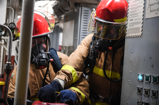 Zwei Feuerwehrleute in Schutzausrüstung und Gasmasken arbeiten an einem Feuerwehrauto, mit einer Tafel und Metallstangen im Hintergrund.