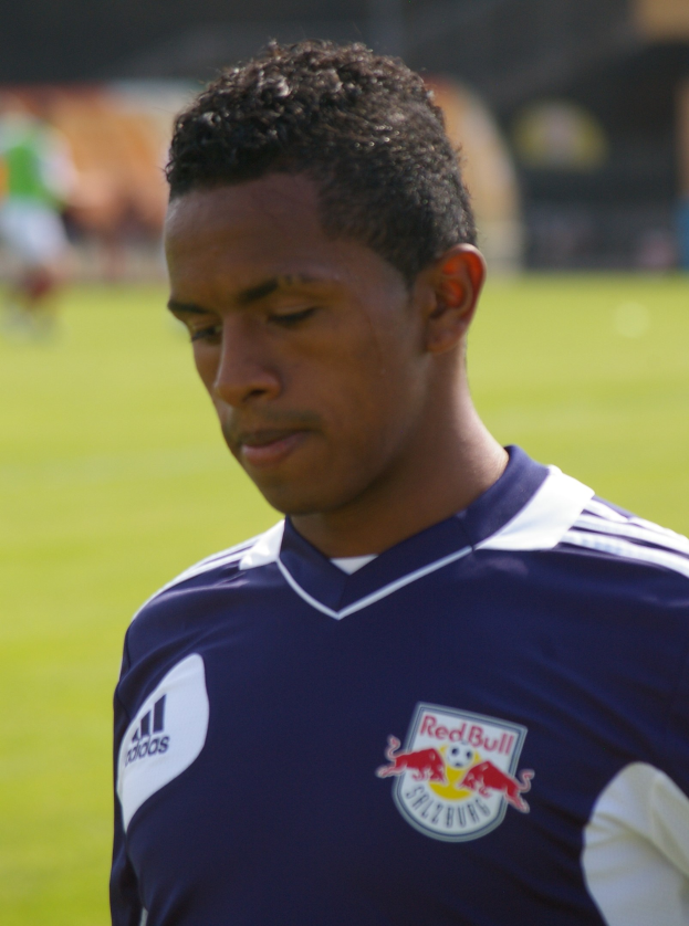 Ein junger Mann in einem blauen T-Shirt steht auf einem Fußballfeld, der als Flügelspieler mit kurzen dunklen Haaren und entschlossener Miene erscheint, vor einem unscharfen Hintergrund.