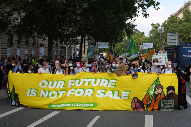 Eine große Gruppe von Menschen, die eine Straße entlanggehen und ein Transparent mit der Aufschrift "Unsere Zukunft ist nicht zu verkaufen" tragen, wobei einige Masken tragen und Taschen tragen, vor dem Hintergrund von Bäumen, Laternenmasten, Gebäuden und einem bewölkten Himmel.