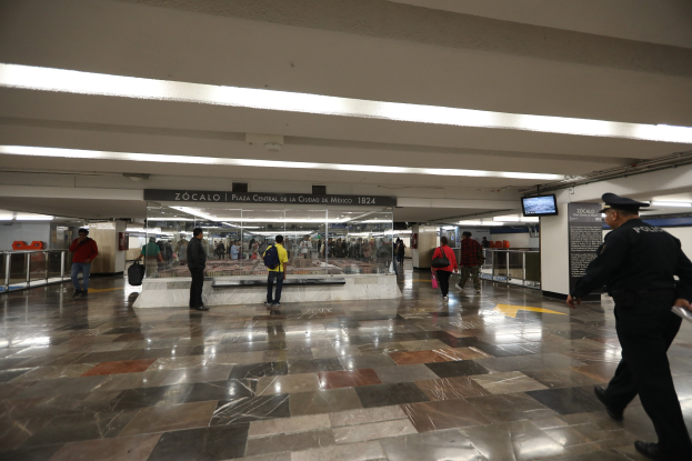 Eine Gruppe von Menschen, die durch eine belebte U-Bahn-Station gehen, mit einem Polizeibeamten im Vordergrund, Leuchttafeln und Bildschirme an der Decke und verschiedenen Geländern und Gegenständen in der Station.