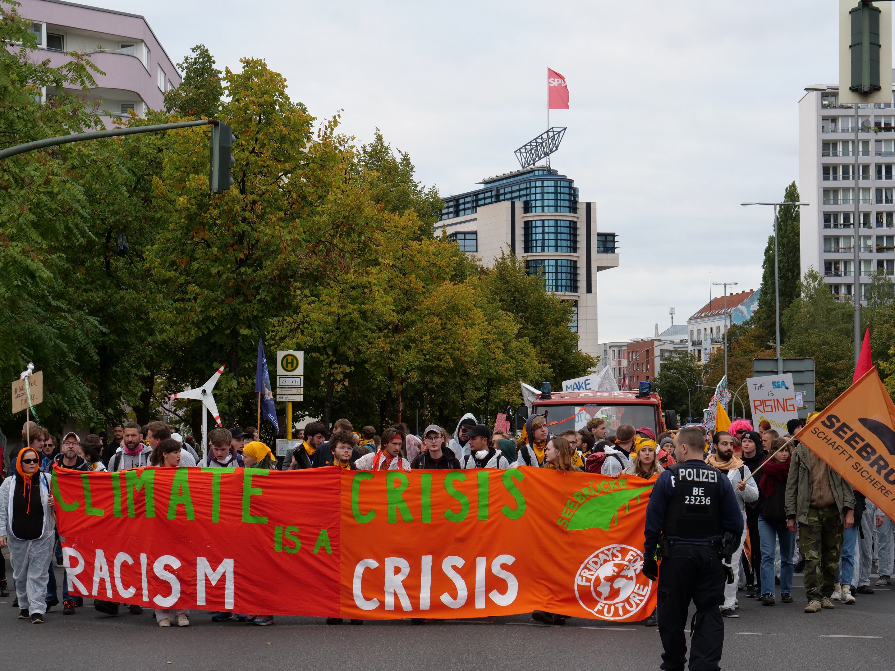 Gruppe von Menschen, die eine Tafel mit der Aufschrift "Klima-Krise ist eine Krise" tragen, auf einer Straße mit Bäumen, Laternen, Schildern, einem geparkten Fahrzeug, Gebäuden und einem klaren blauen Himmel im Hintergrund.