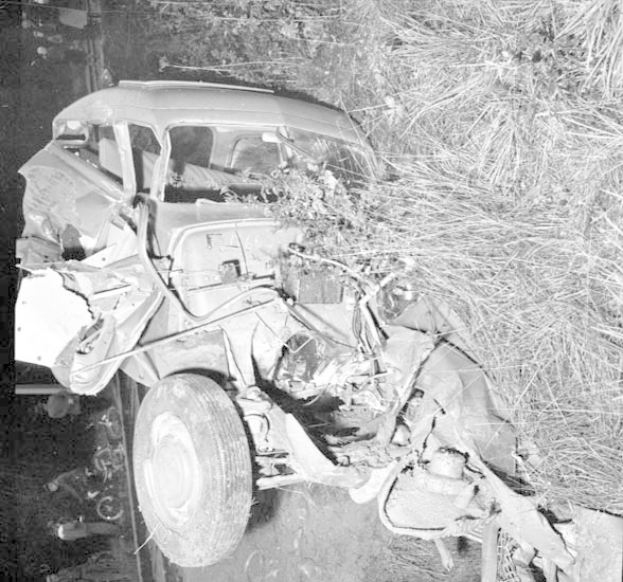Schwarzes und weißes Foto eines schwerbeschädigten Autos mit eingedellter Front und zersplitterten Fenstern am Straßenrand, mit einem Baum und verstreuten Gegenständen im Hintergrund.