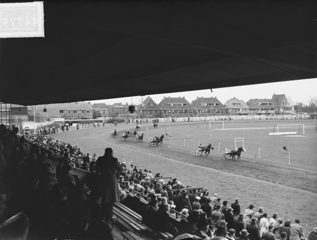 Schwarzes und weißes Foto eines Pferderennens in einer Stadion mit Zuschauern auf Bänken und Jockeys, die Pferde auf der Bahn reiten, mit Gebäuden, Bäumen und Pfählen im Hintergrund unter einem klaren Himmel.