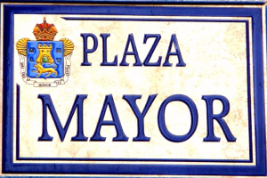 Rechtwinkliges weißes Schild mit schwarzer Schrift "Plaza Mayor" in fetter Schrift, blauem Rand und einem gekrönten Wappen-Logo, an einem Gebäude montiert.