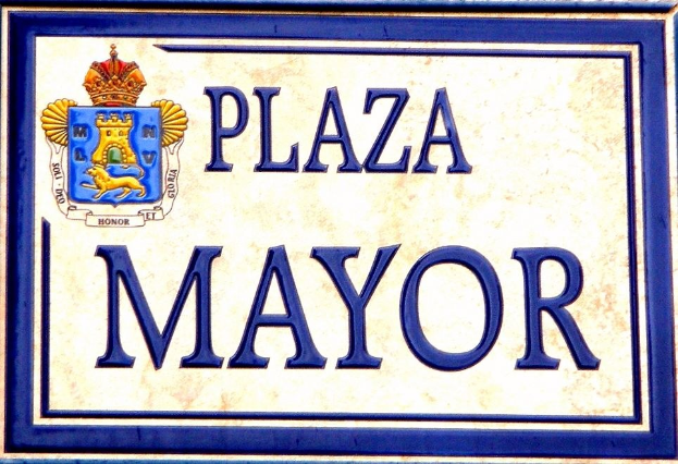 Rechtwinkliges weißes Schild mit schwarzer Schrift "Plaza Mayor" in fetter Schrift, blauem Rand und einem gekrönten Wappen-Logo, an einem Gebäude montiert.