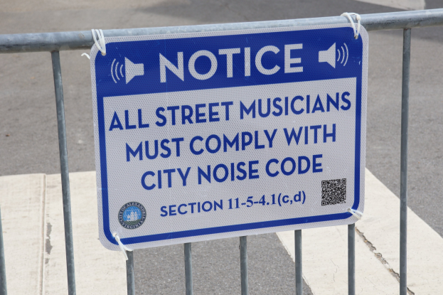 Hinweistafel an einem Zaun mit einem Schild, auf dem "Hinweis: Alle Straßenmusiker müssen die städtische Lärmvorschrift einhalten" steht und eine Straße im Hintergrund.