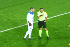 Schiedsrichter in gelbem Trikot und schwarzen Shorts, der mit einem Fußballspieler in weiß-blauer Uniform auf dem Feld spricht, beide tragen Turnschuhe.