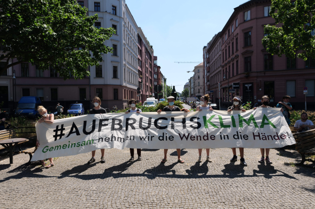 Menschen mit Masken halten ein Transparent mit der Aufschrift 'Aufbruchsklima' während einer Klimaprotest in Berlin, mit Bänken, Tischen, Pflanzen und anderen Personen im Hintergrund.