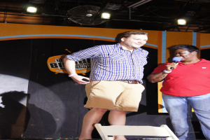 Eine Person in einem roten T-Shirt hält ein Mikrofon und spricht, während eine andere Person neben ihnen lächelt; Fans und Lichter sind oben auf dem Bild zu sehen.