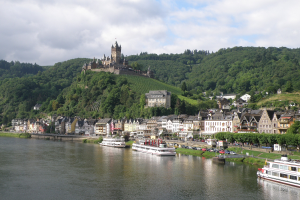 Ein malerischer Blick auf den Rhein in Deutschland, mit einer Burg auf einem Hügel, Booten auf dem Fluss, Fahrzeugen auf der Straße und einem bewölktem Himmel.