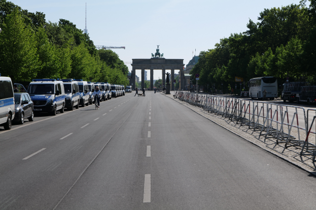 Eine lange Reihe von Polizeiwagen, die auf der Seite einer Straße vor dem Brandenburger Tor in Berlin, Deutschland, geparkt sind, mit Menschen, die Fahrräder fahren und auf der Straße stehen, sowie Absperrungen und Bäume an den Seiten und einem Bogen mit Statuen im Hintergrund.