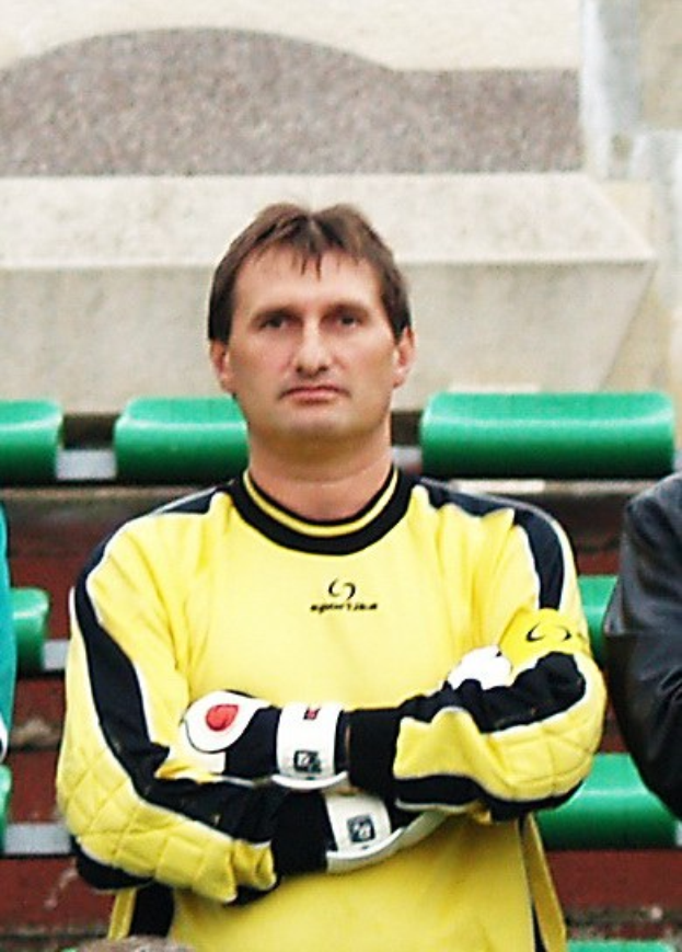 Zwei Männer stehen nebeneinander; der Mann im gelben Trikot, ein Torwart mit konzentrierter Miene, und ein anderer in einer schwarzen Jacke, mit grünen Stühlen und einer Wand im Hintergrund.