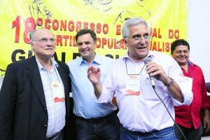 Ein Mann spricht in ein Mikrofon vor einem gelben Vorhang mit der Aufschrift "18-Grad-Kongress o", während andere Männer im Hintergrund stehen.