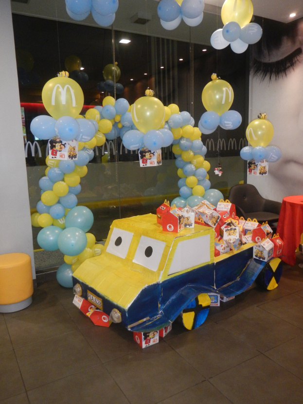 Eine McDonald's-Geburtstagsfeier mit einem gelben und blauen Lastwagen, der mit Ballons und anderen Dekorationen geschmückt ist, einem rot gedeckten Tisch mit Gegenständen, Hockern und einer Glaswand im Hintergrund unter festlichen Deckenlampen.