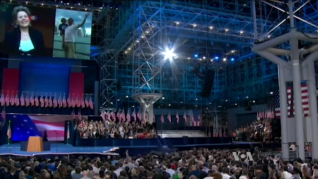 Eine große Menschenmenge sitzt vor einer Bühne mit einem Podium, Mikrofon, Fahnen, einem Bildschirm und Lichtausrüstung und wartet auf eine Präsidentschaftsdebatte.