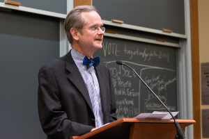 Ein Mann in einem schwarzen Anzug steht an einem Rednerpult und spricht in ein Mikrofon, mit Papieren auf dem Pult und einer Tafel mit Text im Hintergrund.
