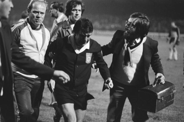 Schwarzes und weißes Bild einer Gruppe von Männern, die auf einem Fußballfeld gehen, einer hält eine Aktentasche, mit einem Fußballspieler in Not in der Mitte.
