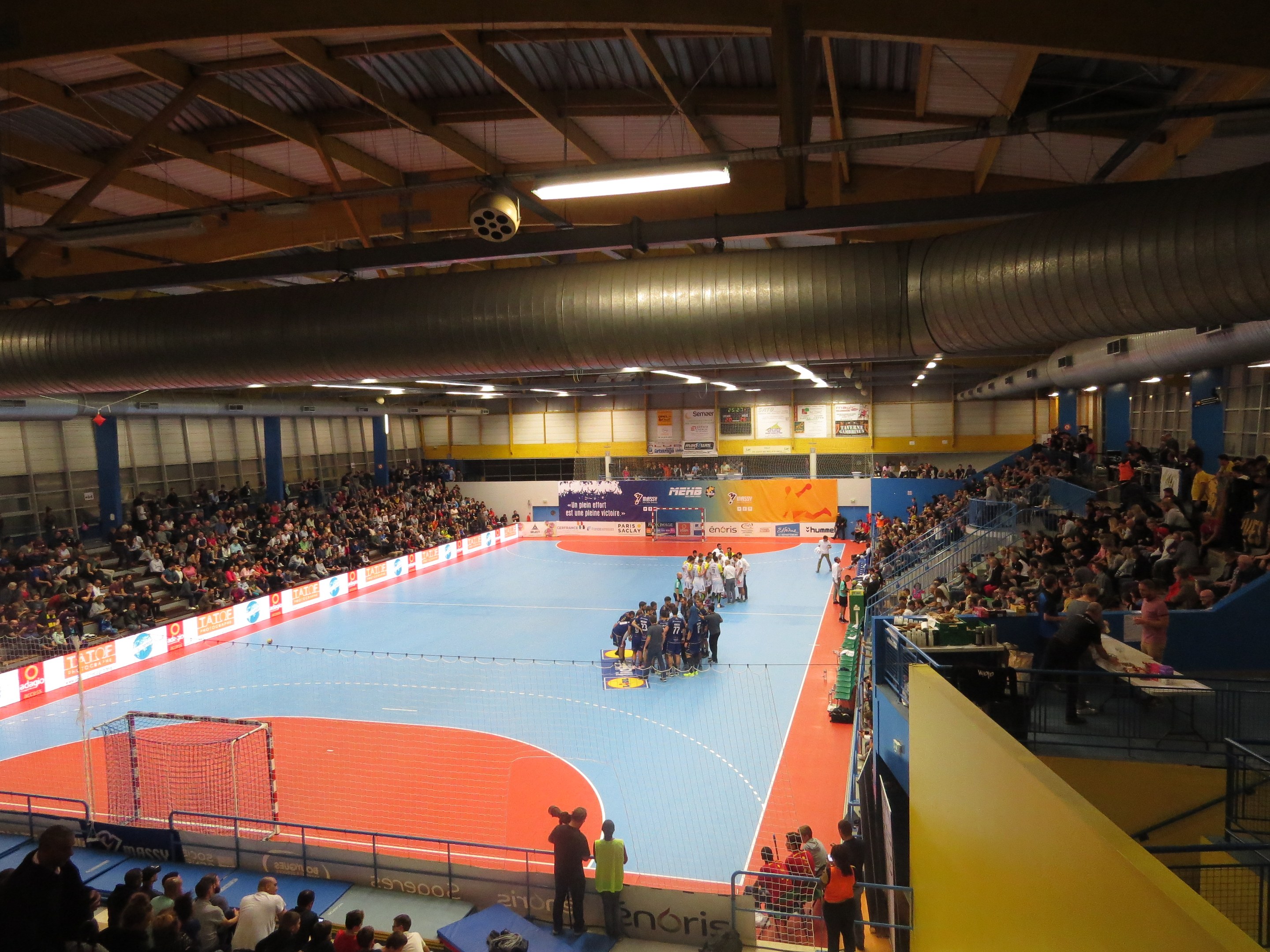 Große Halle mit Zuschauern bei einem Handballspiel, mit Sitzplätzen und Stehplätzen, einem Netz auf der linken Seite, Hintergrundtexttafeln und Deckenbeleuchtung und Rohren.