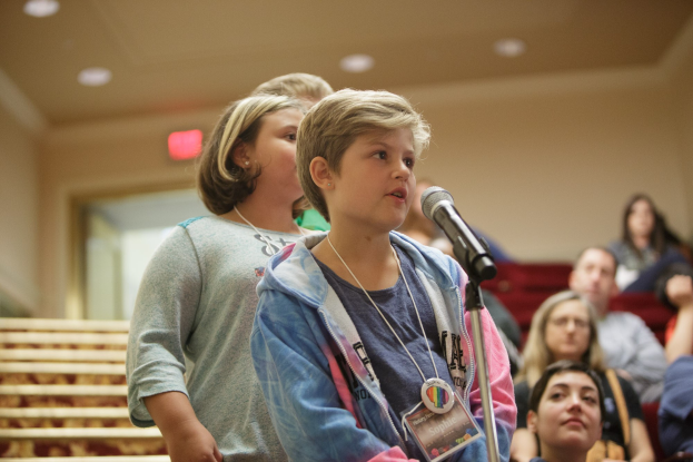 Ein junger Junge steht auf der Bühne und hält ein Mikrofon mit Ständer, schaut auf eine Menge, mit einer Tür in einer Wand und Deckenlampen dahinter.