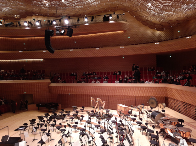 Ein großer Auditorium mit vielen Musikinstrumenten, Stühlen, Ständern mit Papieren und einer Decke, die mit Lichtern beleuchtet ist, zeigt den Hauptsaal der Berliner Philharmonie.