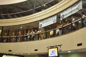 Ein Gebäude mit Menschen an den Fenstern, die nach draußen schauen, zwei Banner an der Scheibe befestigt und ein Fernseher am unteren Bildrand.