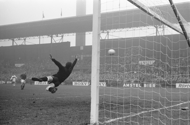 Ein Schwarz-Weiß-Foto eines Fußballspielers bei einer Parade vor dem Tor, mit einem Netz auf der rechten Seite, einer Zuschauermenge im Hintergrund, Tafeln mit Text, einer Brücke und Fahnen mit Stangen.