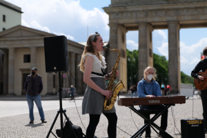 Eine Frau spielt Saxophon vor dem Brandenburger Tor in Berlin, Deutschland, mit einem Pianisten, einem Gitarristen und einem Lautsprecher in der Nähe und Gebäuden, Bäumen und Wolken im Hintergrund.
