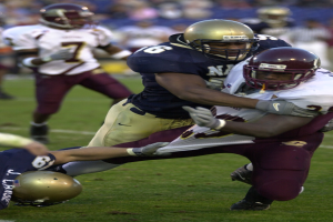 Zwei Fussballspieler in Sportkleidung und Helmen, einer tönt den anderen auf dem Feld, mit anderen Spielern und Zuschauern im Hintergrund.