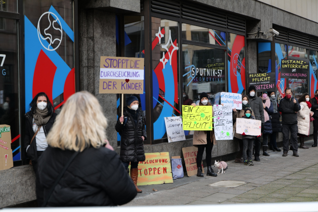 Gruppe von maskierten Menschen mit Schildern vor einem Gebäude mit Glaswänden und -türen, mit Protesttafeln, Kameras und einem Hund im Bild.