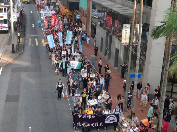 Eine große Gruppe von Menschen marschiert auf einer Straße in Hong Kong, hält Schilder und Transparente, mit Bäumen, gläsernen Gebäuden, Fahrzeugen und Schildern im Hintergrund.
