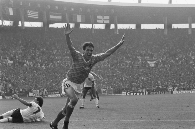 Ein Schwarz-Weiß-Foto eines Fussballspielers, der nach einem Tor jubelt, umgeben von Menschen auf dem Boden, mit Schildern, Fahnen und Pfosten im Hintergrund unter einem klaren Himmel.