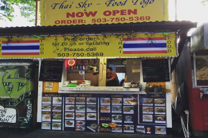 Ein Geschäft mit verschiedenen Gegenständen wie Getränkedosen, Speisekarten, einer Uhr, Gewürzen in Plastikbehältern, Gewürzen, Werbetafeln und Namensschildern unter einem Himmel.