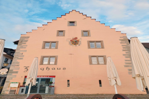 Ein rosafarbenes Gebäude mit einem zentralen Uhrenturm, umgeben von Fenstern, Schirmen, einem Schild, einer Straßenlaterne, Topfpflanzen und einem bewölkten Himmel, repräsentiert München, Deutschland.