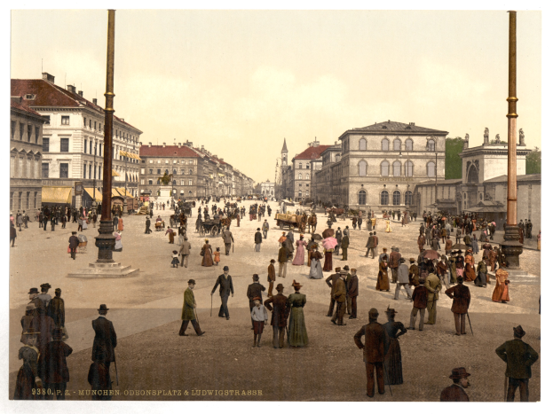 Eine belebte Stadtstraße in München um 1900 mit Menschen, die spazieren gehen, einige mit Schirmen, und einer Kutsche im Vordergrund, Gebäuden, Bäumen und Polen im Hintergrund und Text am unteren Bildrand.