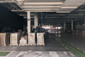 Großes Lagerhaus mit zahlreichen Kartonagen auf Paletten, gestützt von Säulen und beleuchtet von Deckenlampen, mit einem an der Decke angebrachten Schild, das ein Flottenmanagementsystem anzeigt.