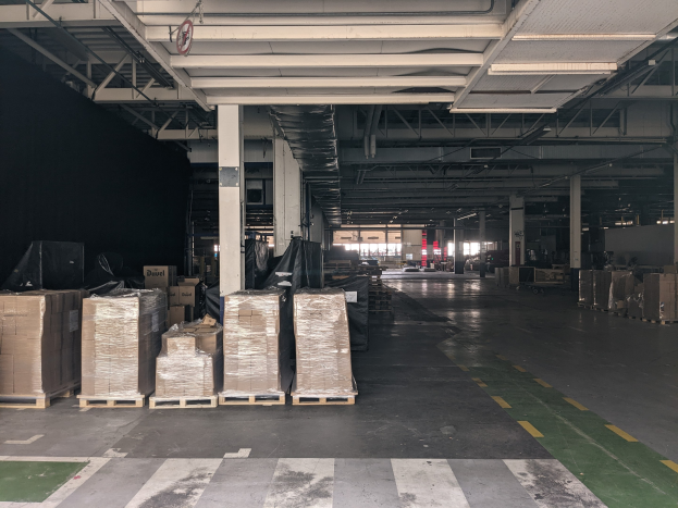 Großes Lagerhaus mit zahlreichen Kartonagen auf Paletten, gestützt von Säulen und beleuchtet von Deckenlampen, mit einem an der Decke angebrachten Schild, das ein Flottenmanagementsystem anzeigt.